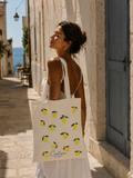 A woman in a white dress stands on a sunlit stone street holding a white tote bag printed with yellow lemons and the text 'Contara STUDIO'.

