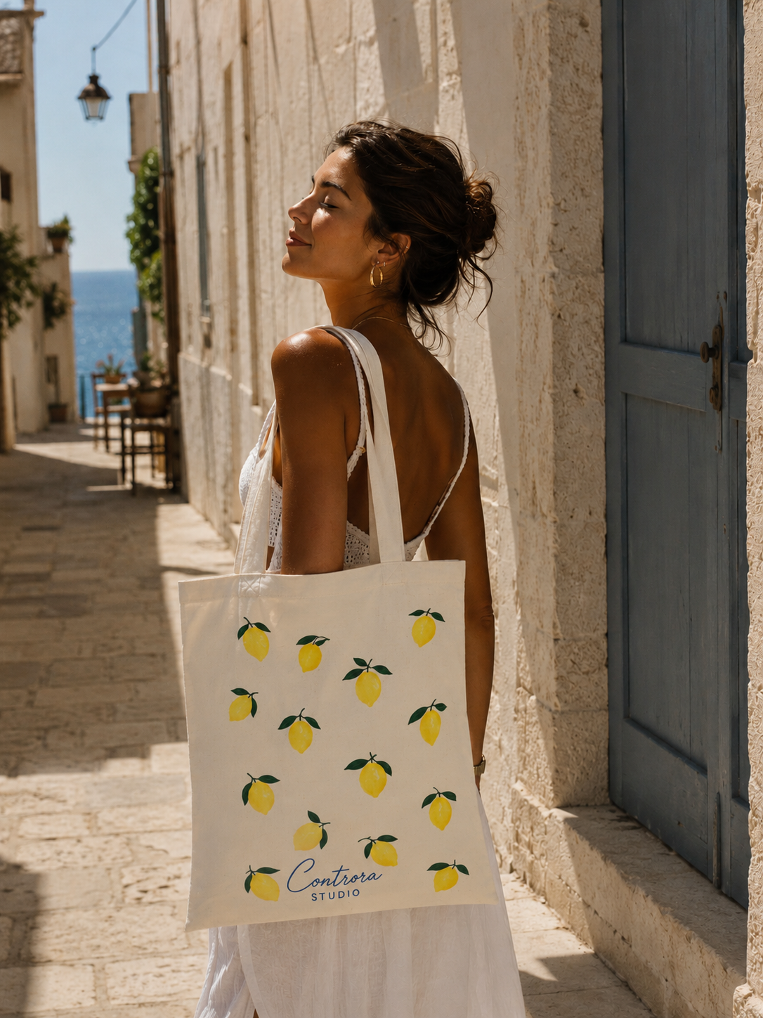 A woman in a white dress stands on a sunlit stone street holding a white tote bag printed with yellow lemons and the text 'Contara STUDIO'.


